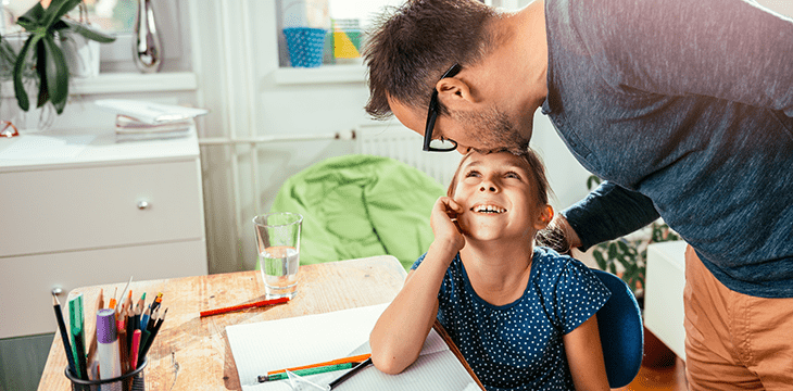 Tochter sitzt an den Hausaufgaben und ihr Vater gibt ihr einen Kuss auf die Stirn.