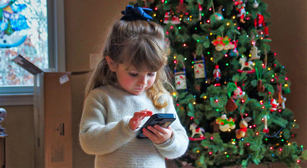 Kleines Mädchen steht vor dem Weihnachtsbaum und tippt auf einem Smartphone herum.
