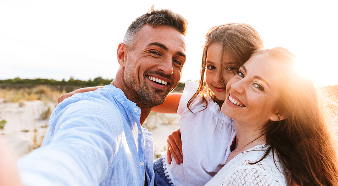 Vater, Mutter und Tochter sind am Strand und machen ein Selfie mit dem Smartphone.