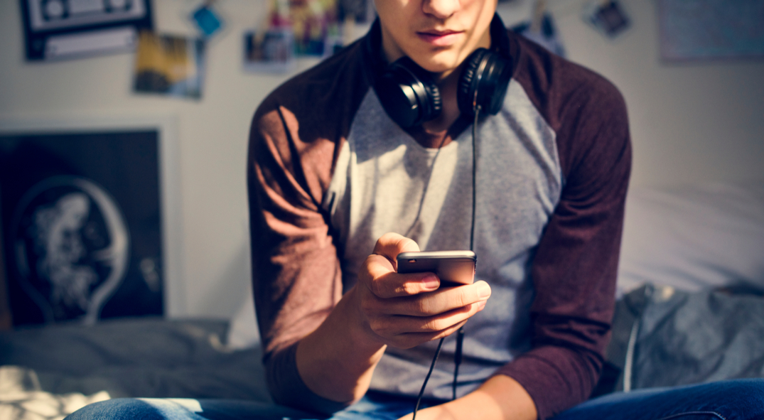 Junge sitzt auf seinem Bett und schaut auf sein Smartphone.