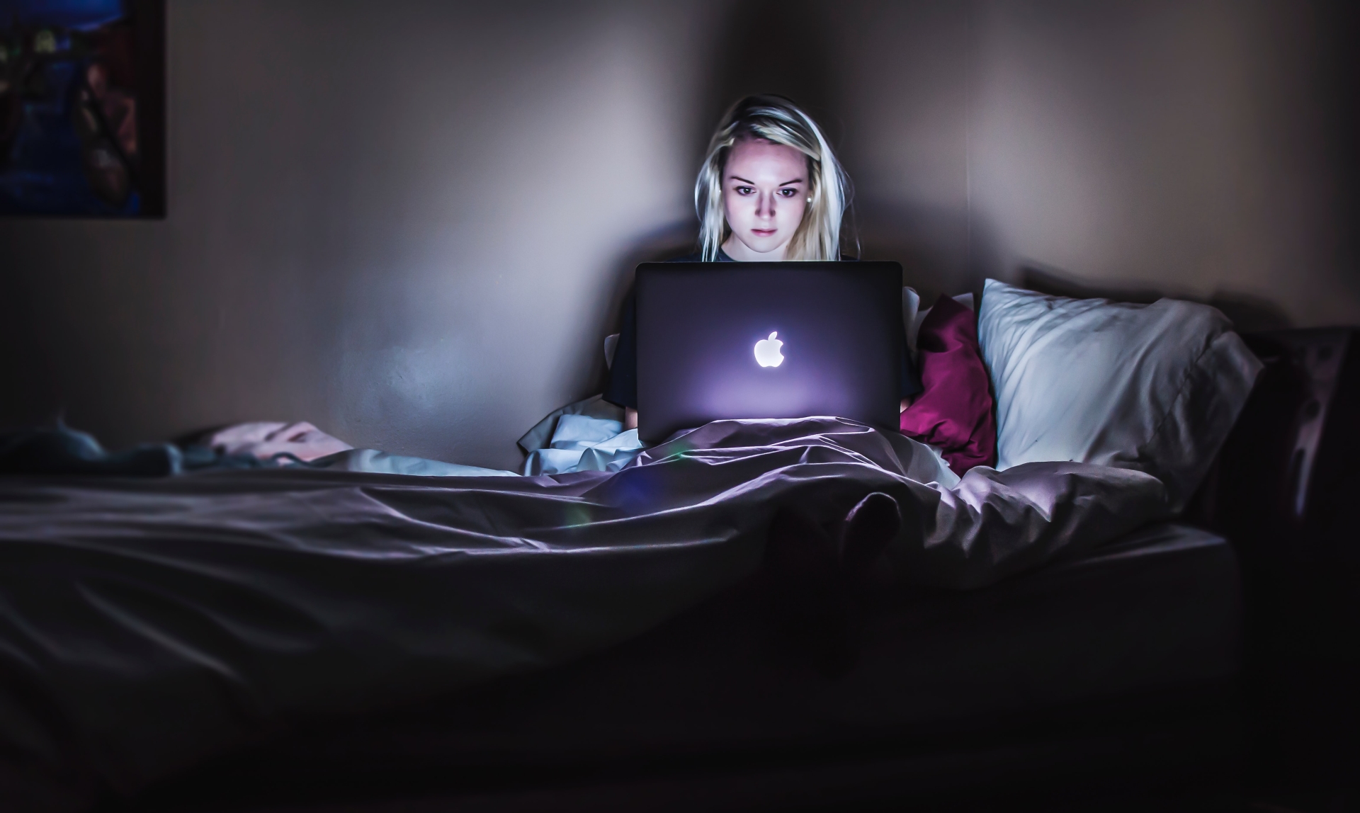 Junge Frau sitzt im Dunkeln auf ihrem Bett. Ihr Gesicht wird vom Laptop angestrahlt, dass sie auf ihrem Schoß hält.