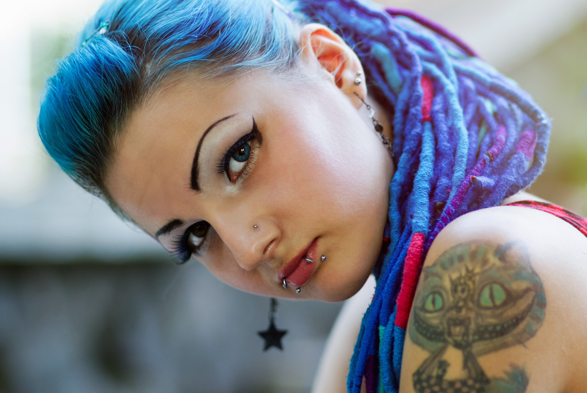 Junge Frau mit blauen Haaren und violetten Rasta-Locken, stark geschminkt mit Tattoo auf der Schulter.