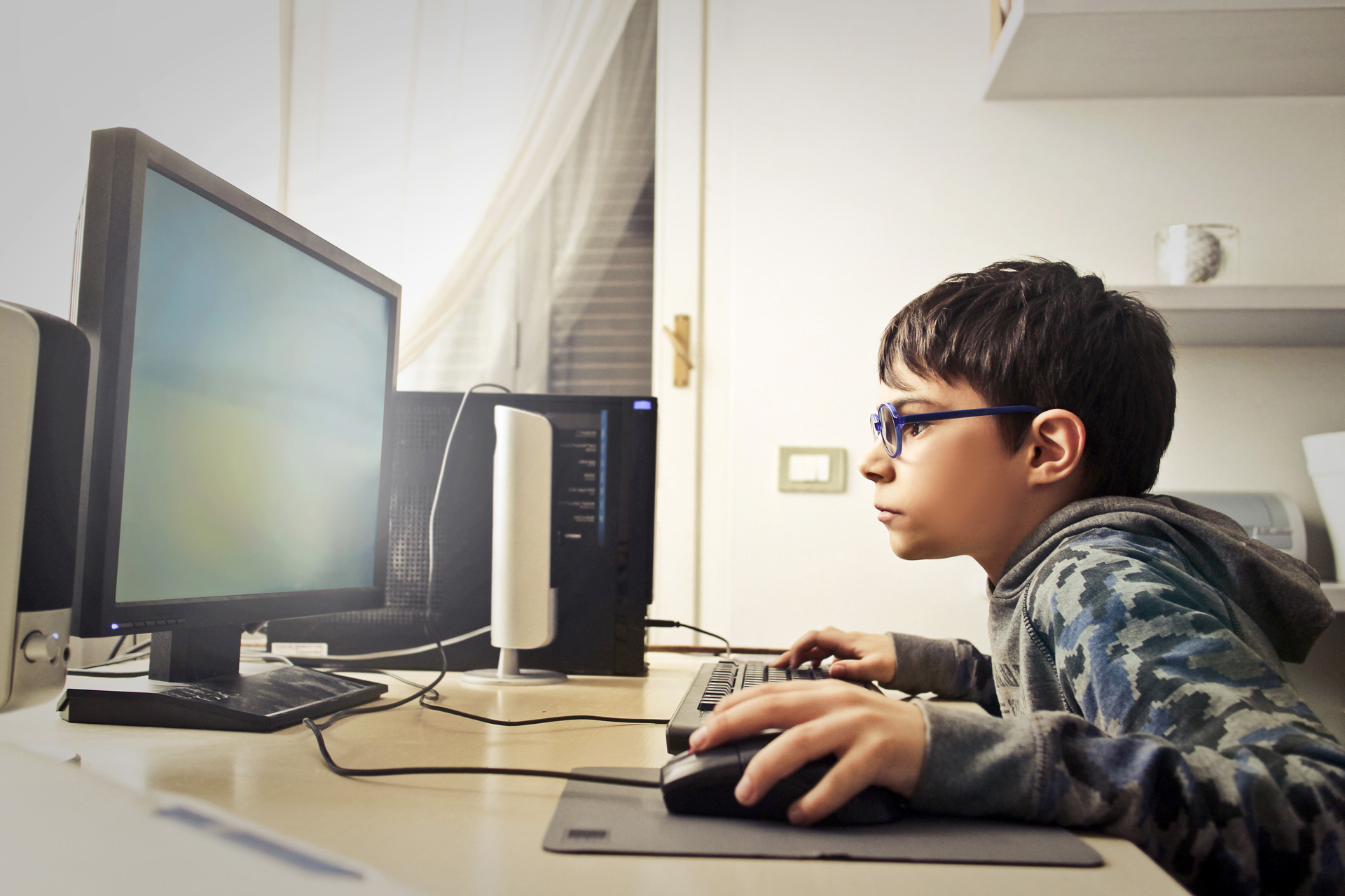 Junge mit Brille sitzt vor einem Computerbildschirm und hat die Maus in seiner Hand.