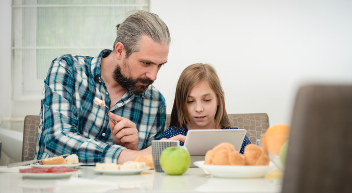 Vater und Tochter sitzen am Esstisch und schauen auf ein Tablet.